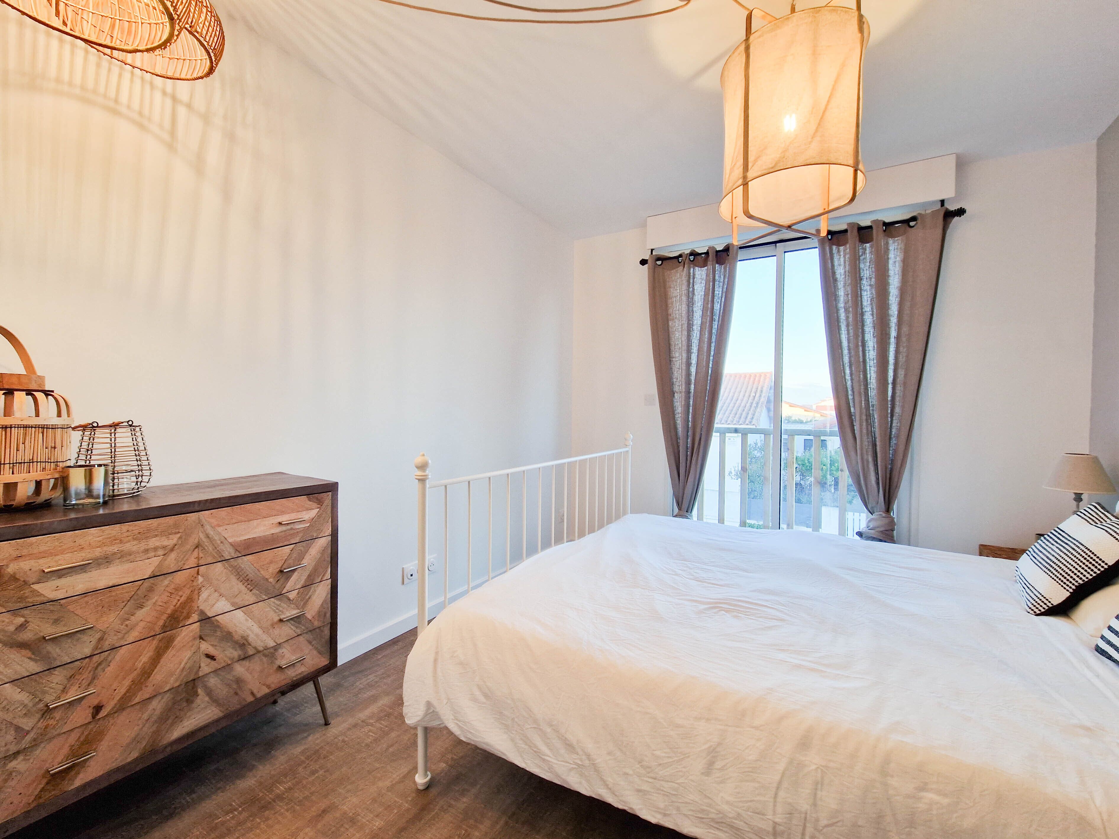 A bedroom with a large bed, a dresser, a window with curtains, and a hanging light fixture.