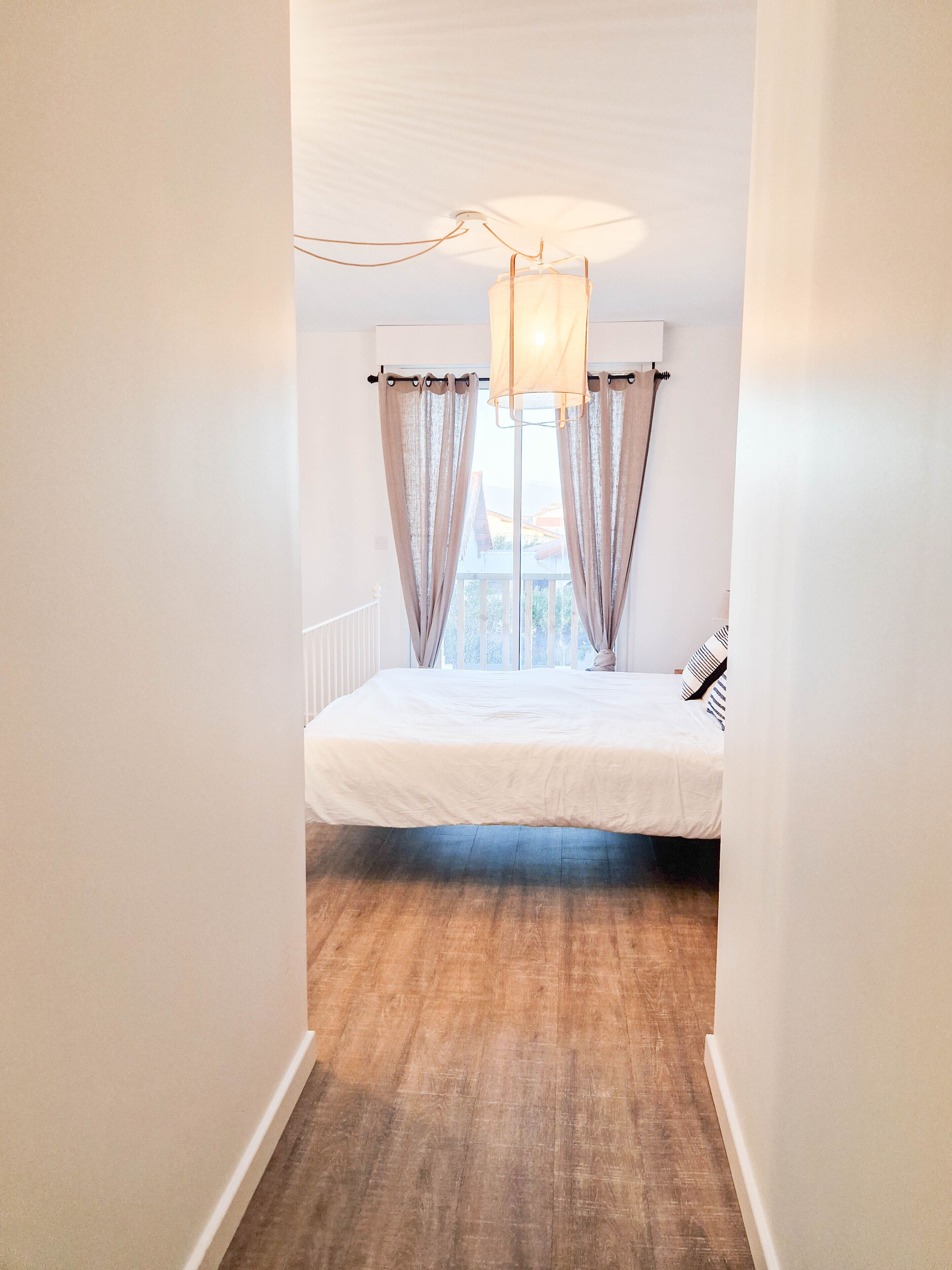 A narrow hallway leading to a bedroom with a bed, curtains, and a hanging light fixture.