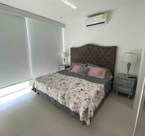 A bedroom with a large bed, two bedside tables, an air conditioning unit, and a sliding glass door.