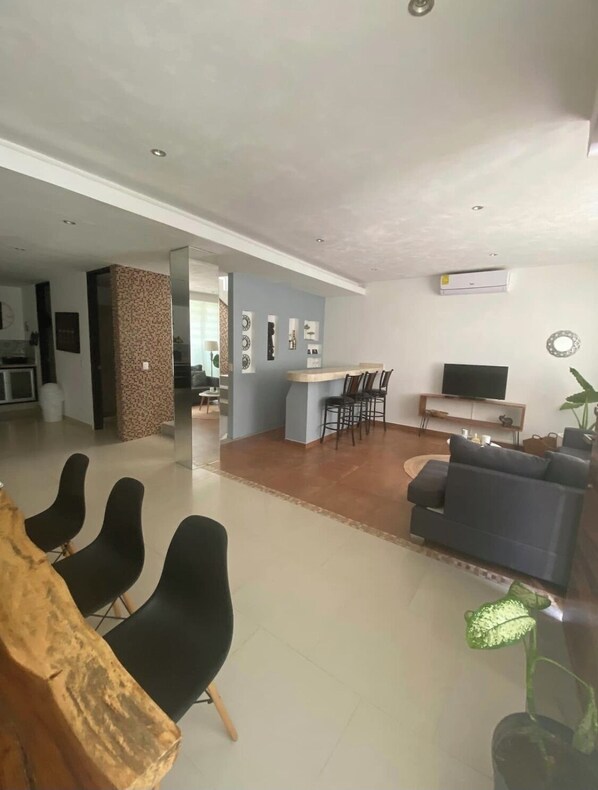 A modern living room with a sofa, chairs, a coffee table, and a TV. There is a kitchen area in the background with a dining table and chairs.