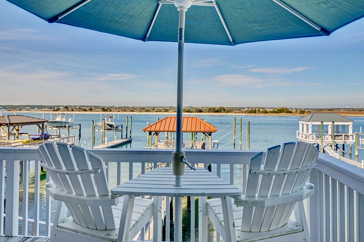A deck with two white Adirondack chairs, a table, and a blue umbrella.
