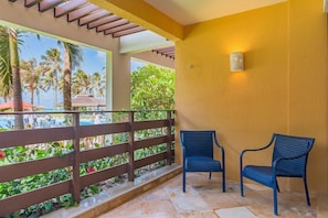 Un balcon avec deux fauteuils en osier, une clôture en bois et une vue sur un espace piscine agrémenté de palmiers.