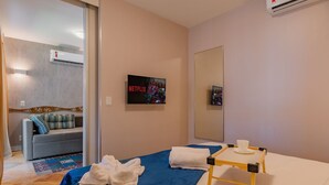 A hotel room with a bed, a small table with a cup, a wall-mounted TV displaying Netflix, a sofa with a cushion, and an air conditioning unit. - Apart no Beach Park Suítes por Carpediem (Aquiraz)