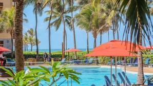 A pool area with lounge chairs and red umbrellas, surrounded by palm trees and a view of the ocean. - Apart no Beach Park Suítes por Carpediem (Aquiraz)