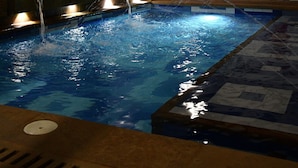 Ein Schwimmbecken mit Wasserstrahlmassagen, einer gefliesten Terrasse und einer weißen Wand mit einem Ziegeldach.