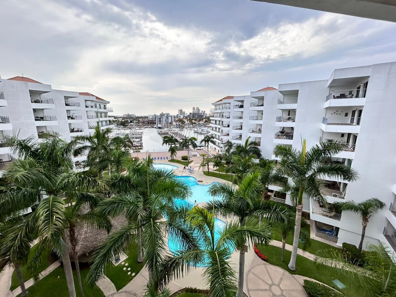 A resort with a central swimming pool surrounded by palm trees and white buildings with balconies.