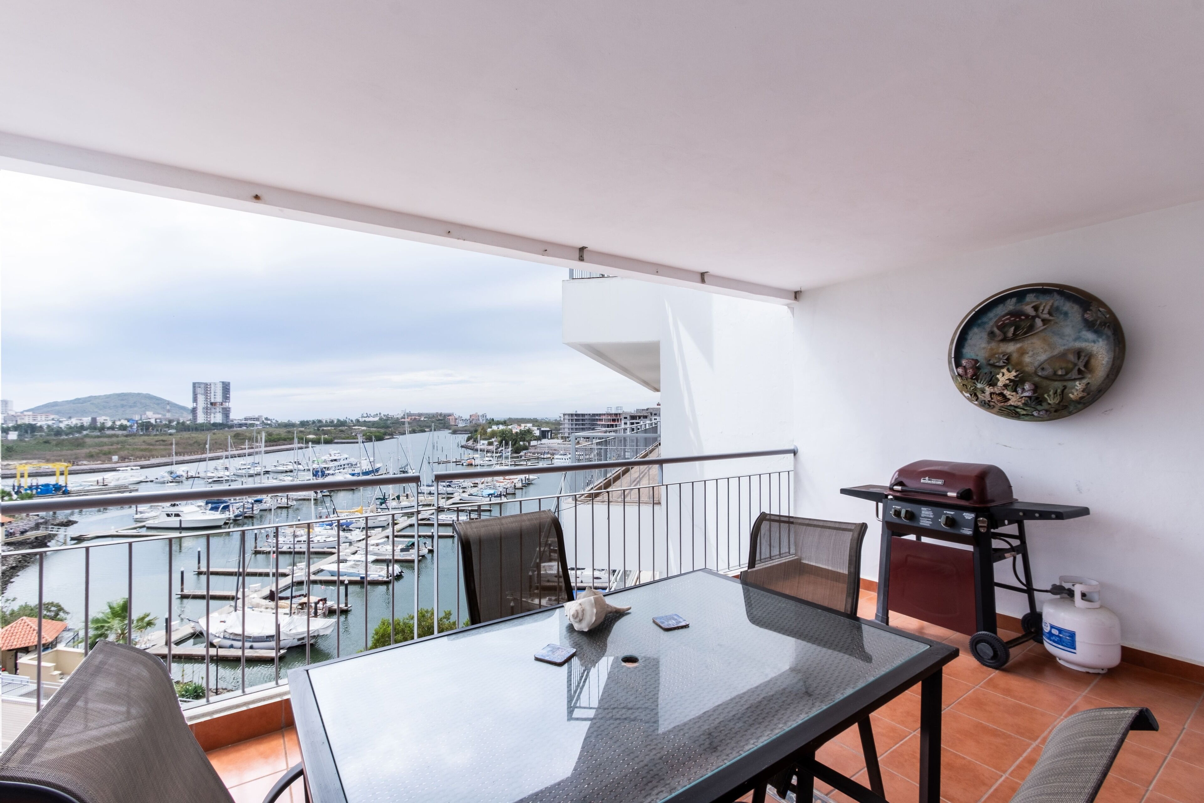 A balcony with a glass table, chairs, a grill, and a view of a marina and cityscape.