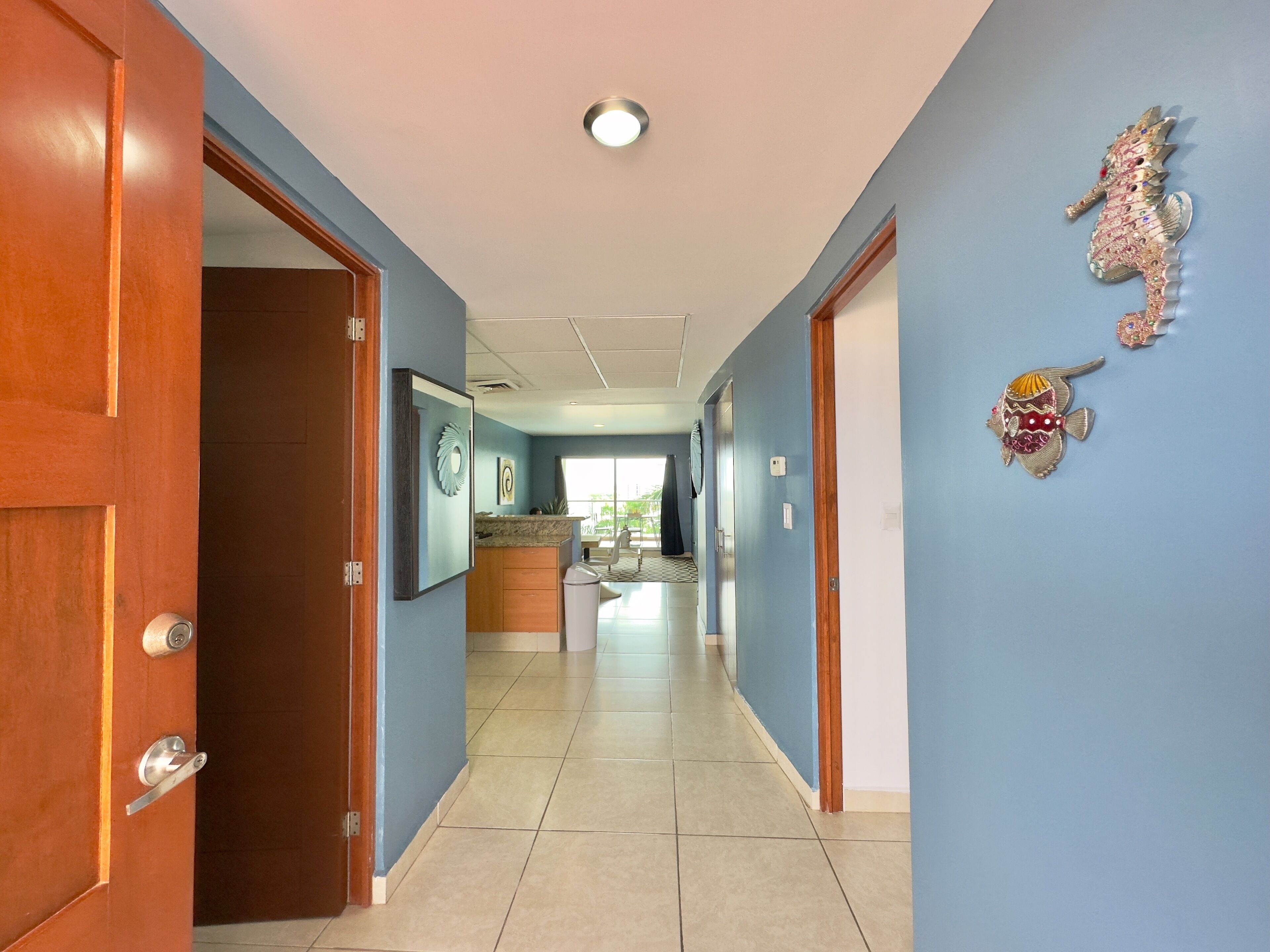 Un couloir avec une porte en bois sur la gauche, un mur bleu décoré de motifs de poissons et, au fond, une vue sur une pièce comprenant un coin cuisine.