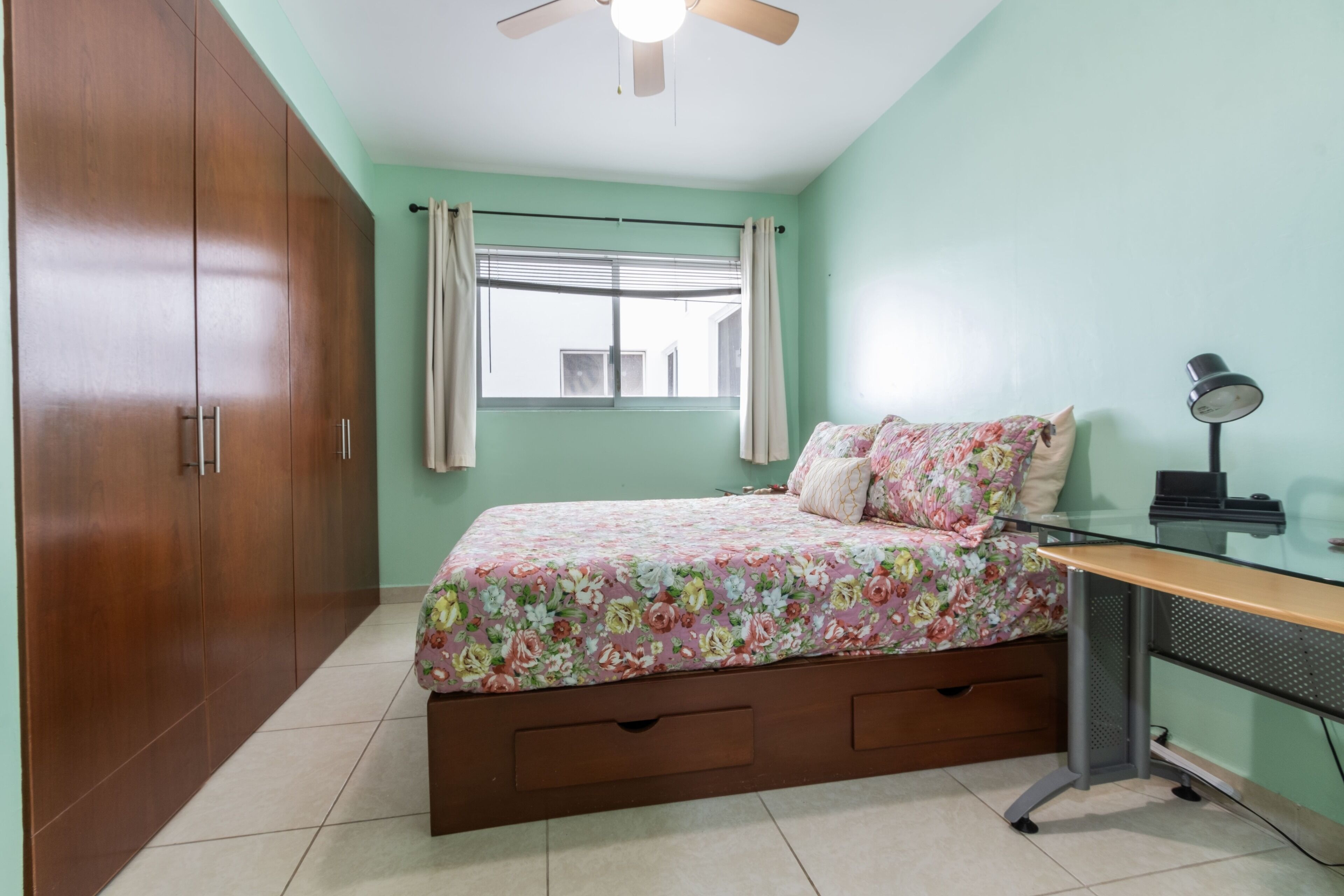 A bedroom with a bed, a desk, a ceiling fan, and a window with curtains.