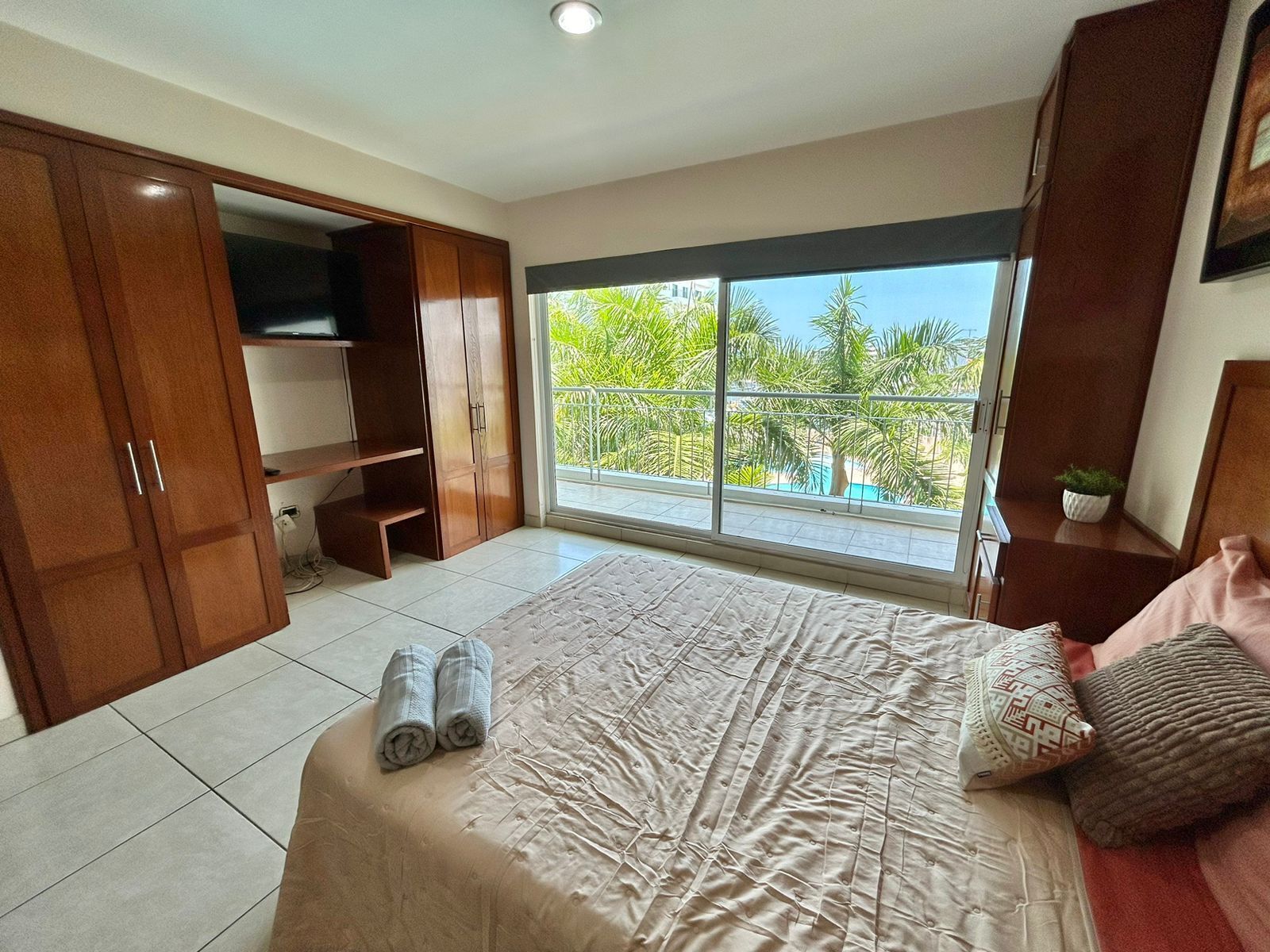 A bedroom with a large bed, a wardrobe, a TV, and a balcony with a view of greenery.