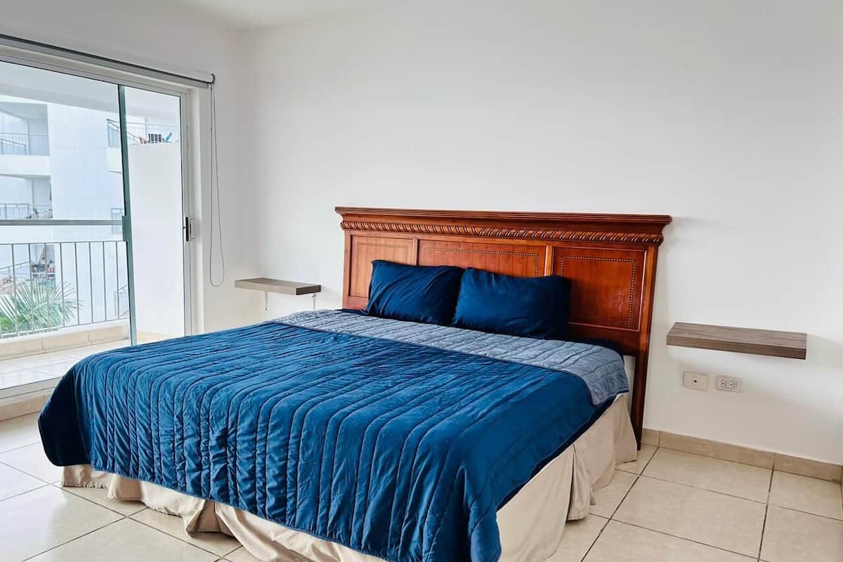 A bedroom with a large bed, a wooden headboard, a window with a view, and a bedside table.