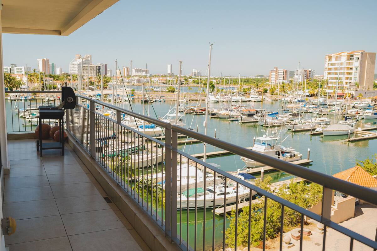 A balcony with a view of a marina filled with boats, a cityscape in the background, and a tiled floor.