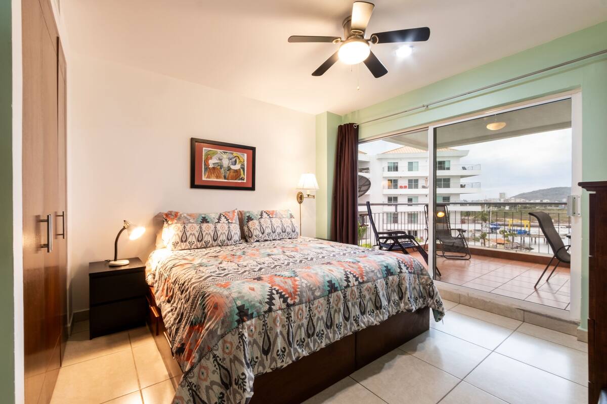 A bedroom with a bed, nightstand, lamp, and a view of the balcony.