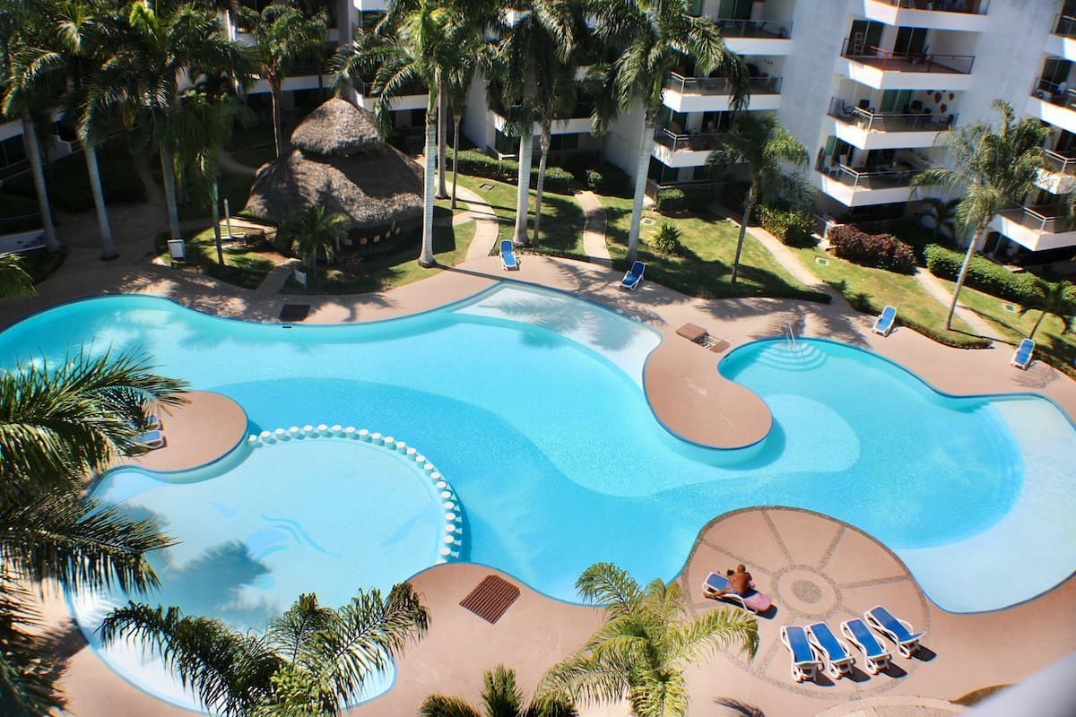A large, irregularly shaped swimming pool with multiple levels and lounge chairs around it, surrounded by palm trees and a building with balconies.