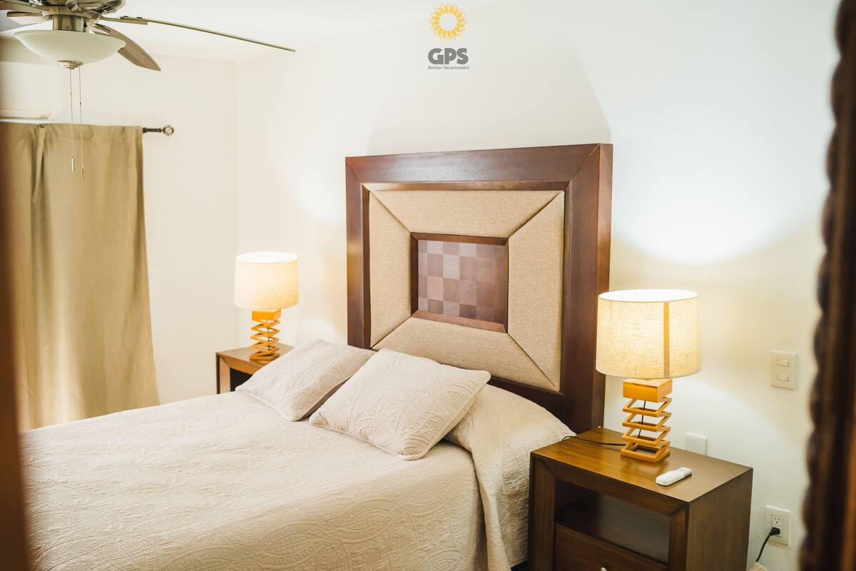 Quarto de hotel com cama, duas mesas de cabeceira com luminárias, um ventilador de teto e um logotipo fixado na parede.