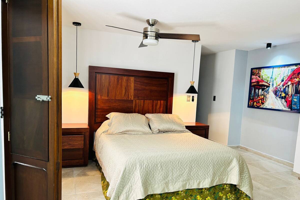 Quarto com cabeceira de madeira, cama com cobertura acolchoada, duas luminárias de cabeceira, ventilador de teto e uma pintura na parede.