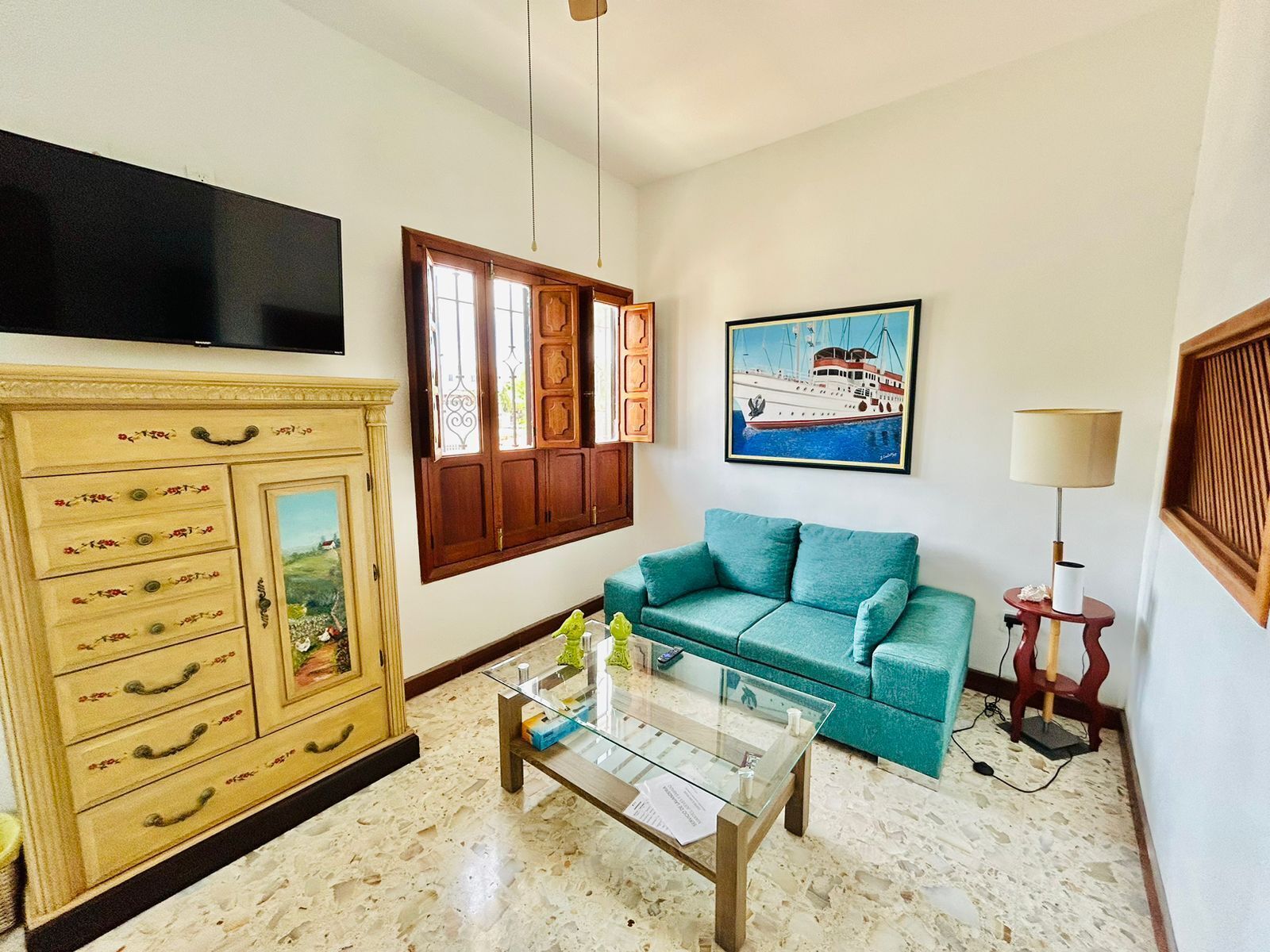 A living room with a teal sofa, a glass coffee table, a wooden cabinet, a painting of a boat, and a television on the wall.