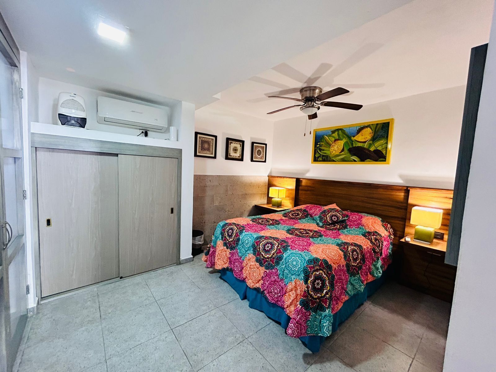 A bedroom with a bed, a ceiling fan, a painting, and a nightstand with a lamp.