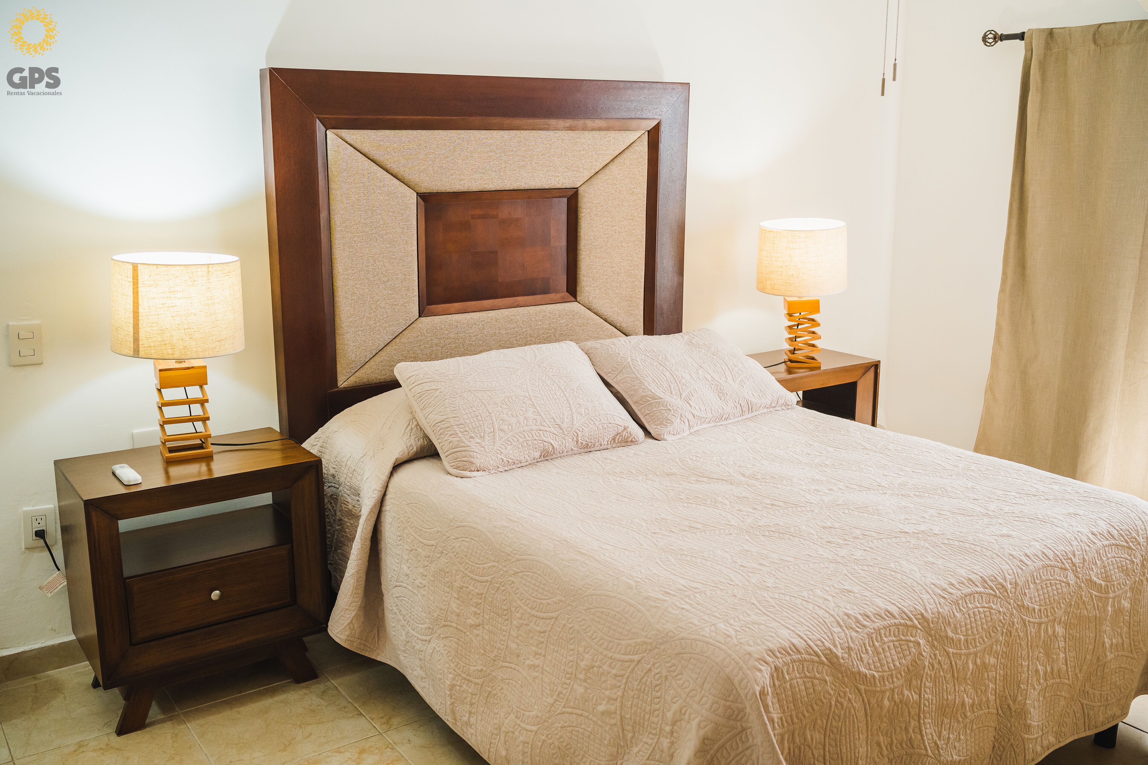 A neatly made bed with a wooden headboard, two bedside tables with lamps, and a nightstand with a drawer.