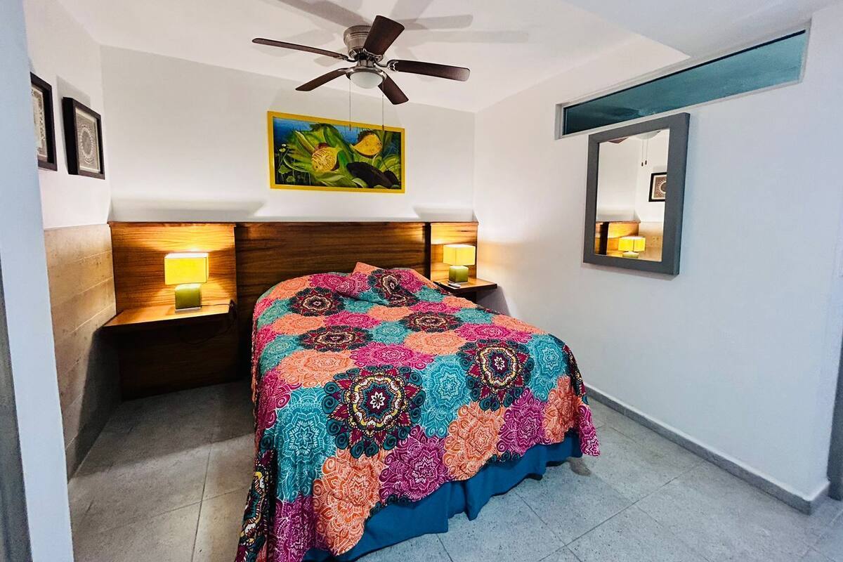 Quarto com cama, duas luminárias de cabeceira, ventilador de teto, um quadro na parede e um espelho.