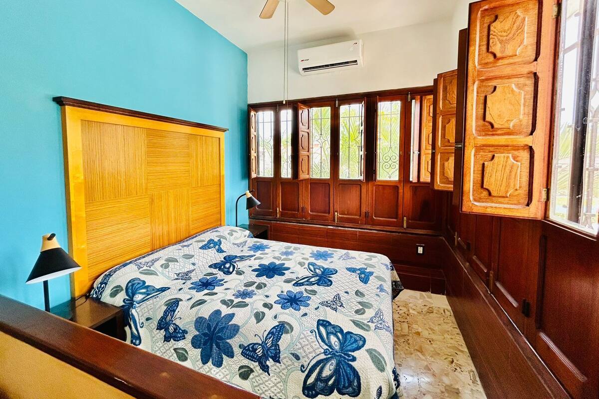 Quarto com cabeceira de madeira, cama com colcha florida, ventilador de teto e janela com persianas de madeira.
