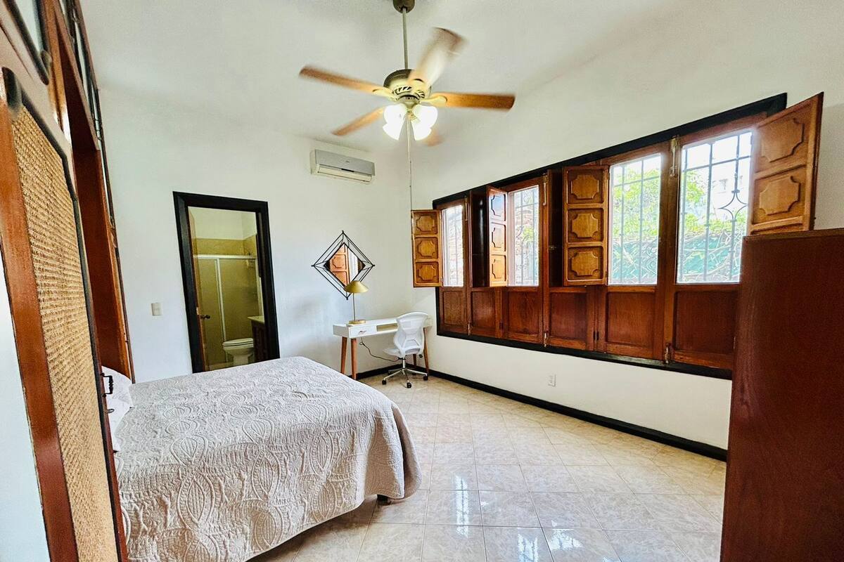 Um quarto com uma cama, uma escrivaninha, uma cadeira, um ventilador de teto e uma janela com persianas de madeira.