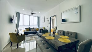 Un salón moderno con un sofá gris, cojines amarillos, una mesa de comedor con manteles individuales amarillos y un espejo grande en la pared.