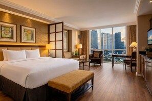 A hotel room with a large bed, a desk, a chair, and a view of the cityscape through the windows.