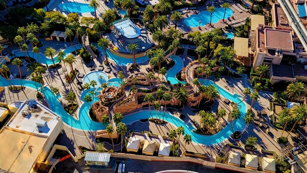 Vista aérea de un complejo turístico con varias piscinas, palmeras y una formación rocosa central.