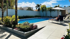 Una piscina con plataforma de azulejos, rodeada de palmeras y plantas, y una persona relajándose en una tumbona.