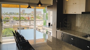 Una cocina moderna con encimeras de granito, armarios negros e iluminación colgante. Se ve un comedor con mesa y sillas a través de un amplio ventanal con vistas a la ciudad.