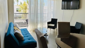 Una sala de estar con un sofá azul, un sillón marrón, una mesa de centro de vidrio y un televisor montado en la pared.