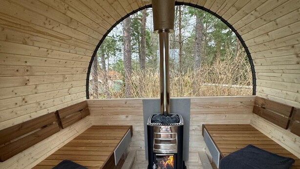 Sala de sauna con bancos de madera, una unidad de calefacción central y vistas a árboles a través de un techo de madera semicircular.