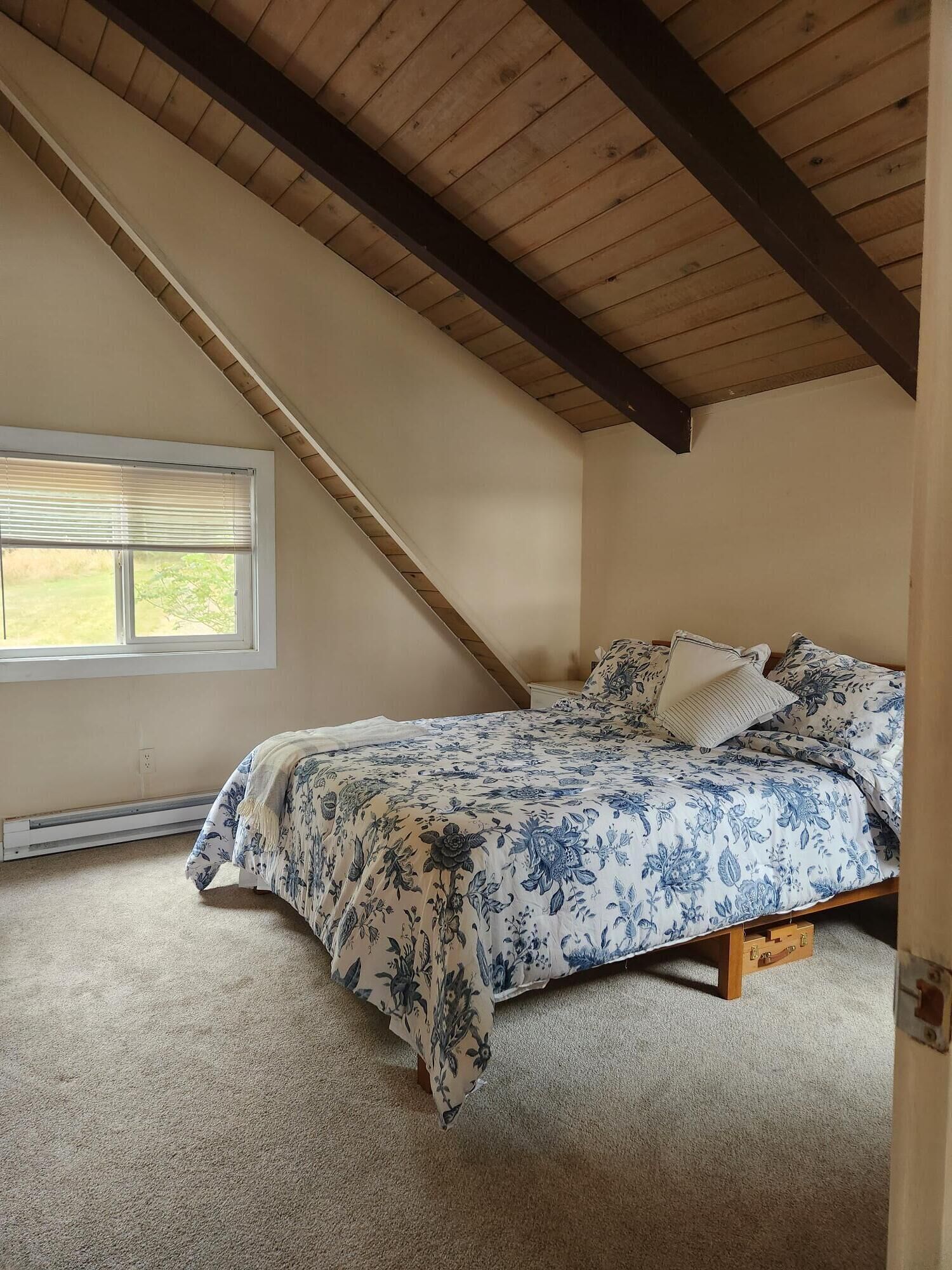 A bedroom with a sloped ceiling, a bed with floral bedding, a window with blinds, and a carpeted floor.