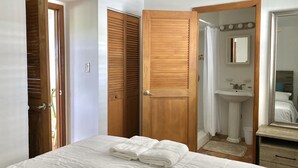 Quarto com cama, portas de madeira, banheiro com pia e espelho, e um tapete pequeno.