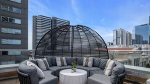 A rooftop terrace with a sectional sofa and a round table, overlooking a cityscape with modern buildings.
