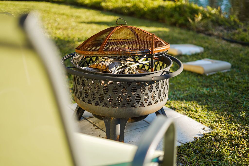 A metal fire pit with a wire mesh cover, surrounded by grass and a concrete base.