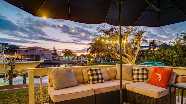 A covered outdoor seating area with a sofa and umbrella, overlooking a marina with boats and a sunset sky. - Manatee Cove: Private Oasis with Wildlife, Hot Tub & Fun Game Room! (Bradenton)