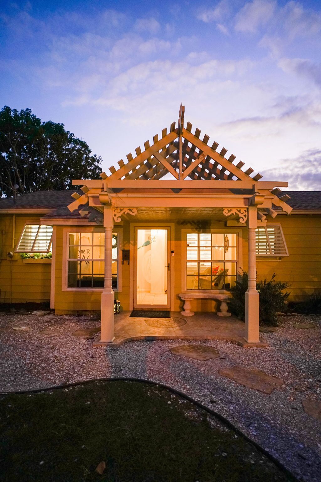 A yellow building with a wooden pergola entrance, a bench, and a gravel driveway.
