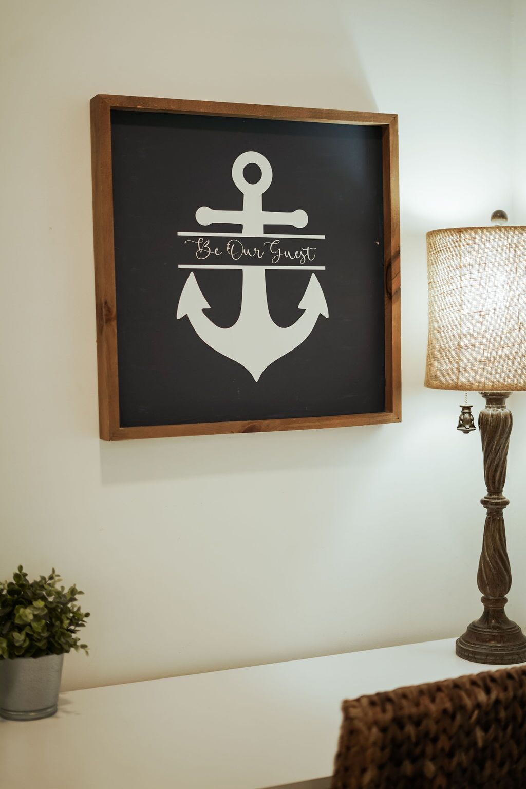 Un adorno enmarcado con forma de ancla y el texto 'Be Our Guest' en una pared.