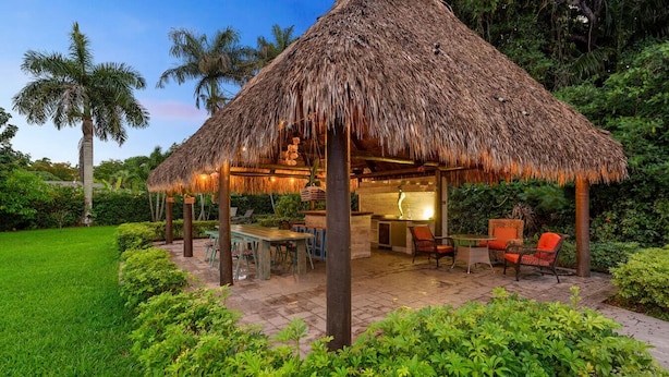 A thatched-roof outdoor dining area with tables and chairs, surrounded by lush greenery and palm trees. - Huge Ultra Estate on an Acre! Night Club, Gameroom, Hot-Tub. Pool, Tiki (North Miami Beach)