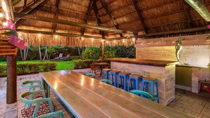 An outdoor bar area with a thatched roof, wooden bar counter, stools, and a long table. - Huge Ultra Estate on an Acre! Night Club, Gameroom, Hot-Tub. Pool, Tiki (North Miami Beach)
