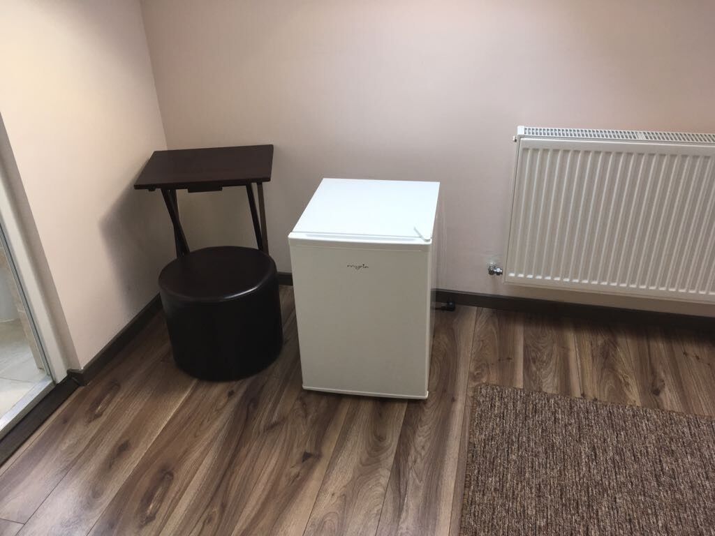A compact room with a small refrigerator, a black trash bin, a table, and a radiator.