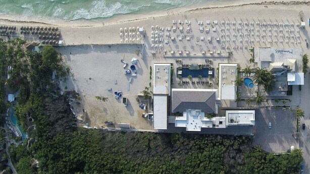 An aerial view of a coastal hotel with a beach area, multiple swimming pools, and a parking lot.