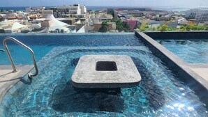 A rooftop infinity pool with a stone hot tub, clear blue water, and a city view in the background.