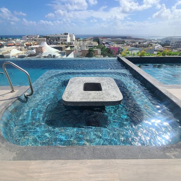 A rooftop infinity pool with a stone hot tub, clear blue water, and a city view in the background.
