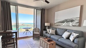 Ein großzügiger Wohnbereich mit grauem Sofa, Couchtisch und Sessel aus Weidengeflecht, Deckenventilator und einem großen Fenster mit Blick ins Freie.