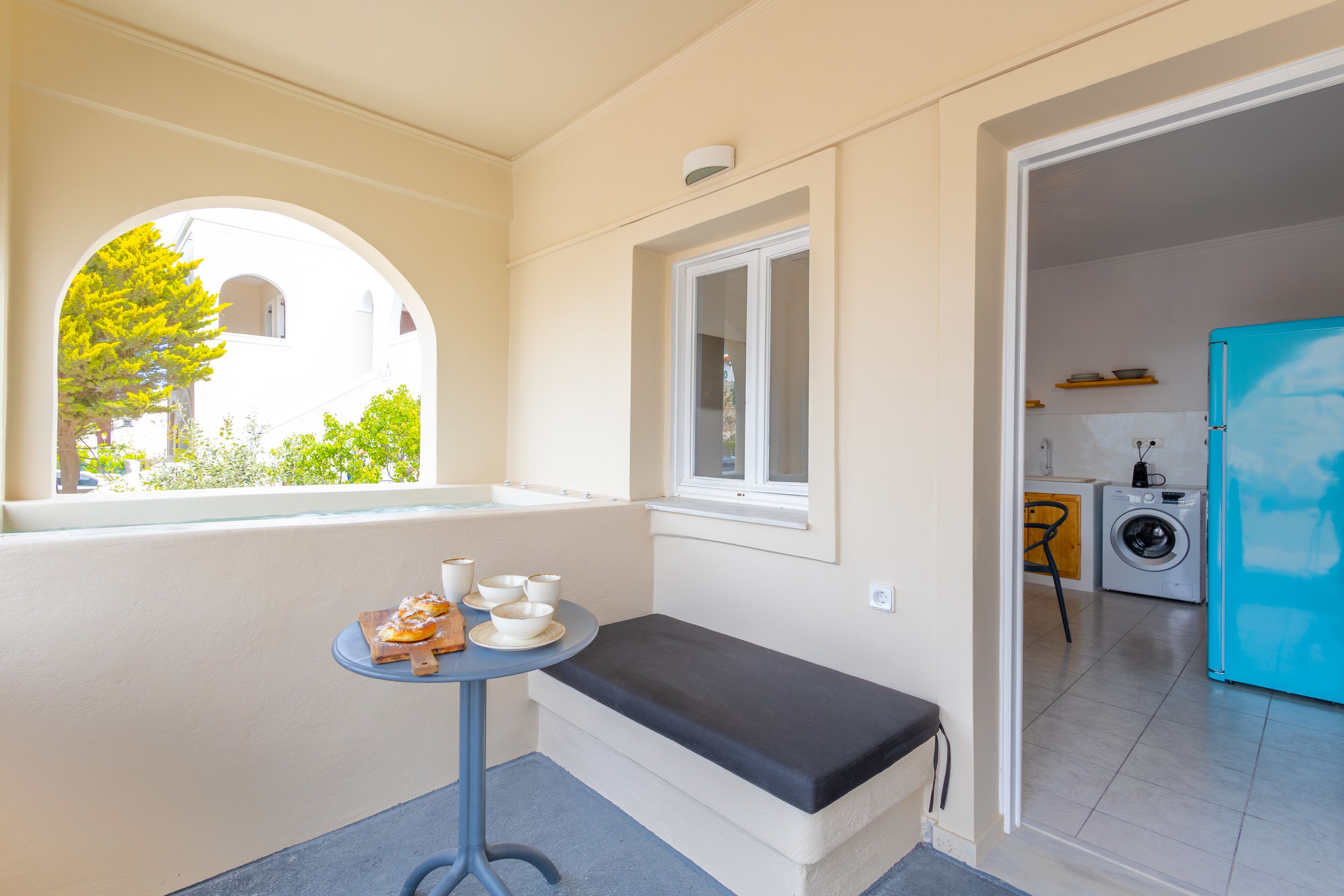 A compact room with a small table, a bench, a window with a view of greenery, and a kitchen area with a washing machine and a blue refrigerator.