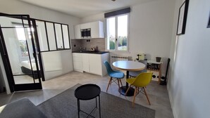 A modern kitchen with white cabinets, a built-in oven, and a window. There is a dining area with a round table and two chairs, one blue and one yellow. - Le repaire de Barbizon (Barbizon)