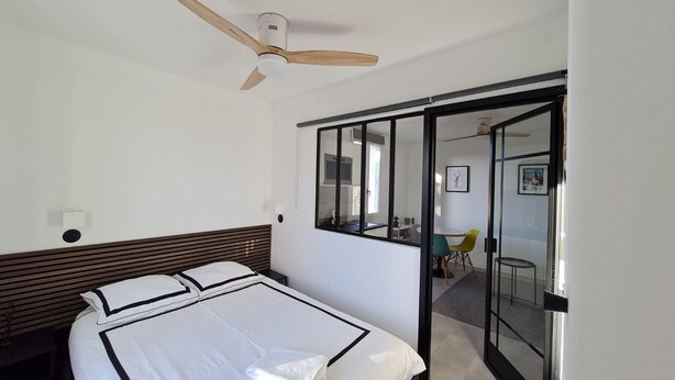 A bedroom with a bed, a ceiling fan, a sliding glass door leading to a dining area, and a window. - Le repaire de Barbizon (Barbizon)